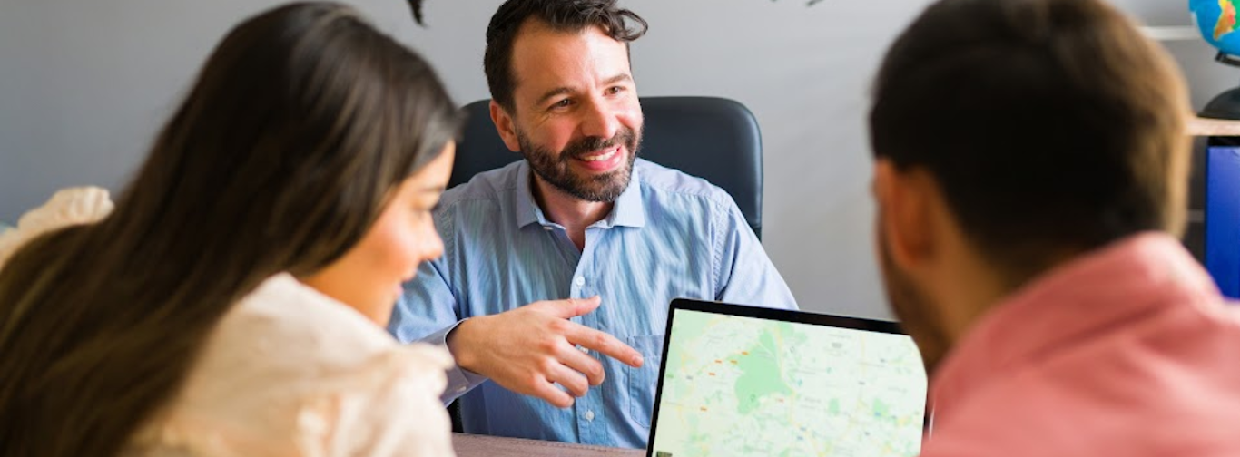 Travel agent consulting clients using travel agency software with digital maps and booking tools.